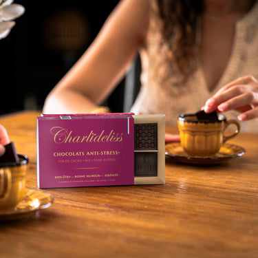 Femme brune trempant un carré de chocolat Charlidéliss dans sa tasse de café en face d'une autre personne qui fait de même. Le coffret de chocolat est posé au milieu et on voit très clairement indiqué ses bienfaits anti-stress naturels puissants.